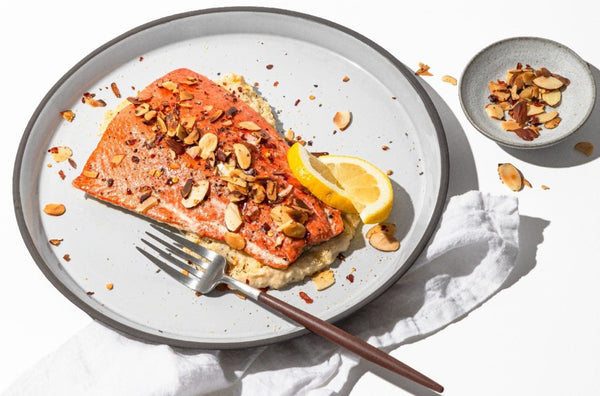 Cooked wild Alaskan king salmon fillet topped with toasted sliced almonds, cracked pepper, and a lemon wedge on a gray plate with a fork