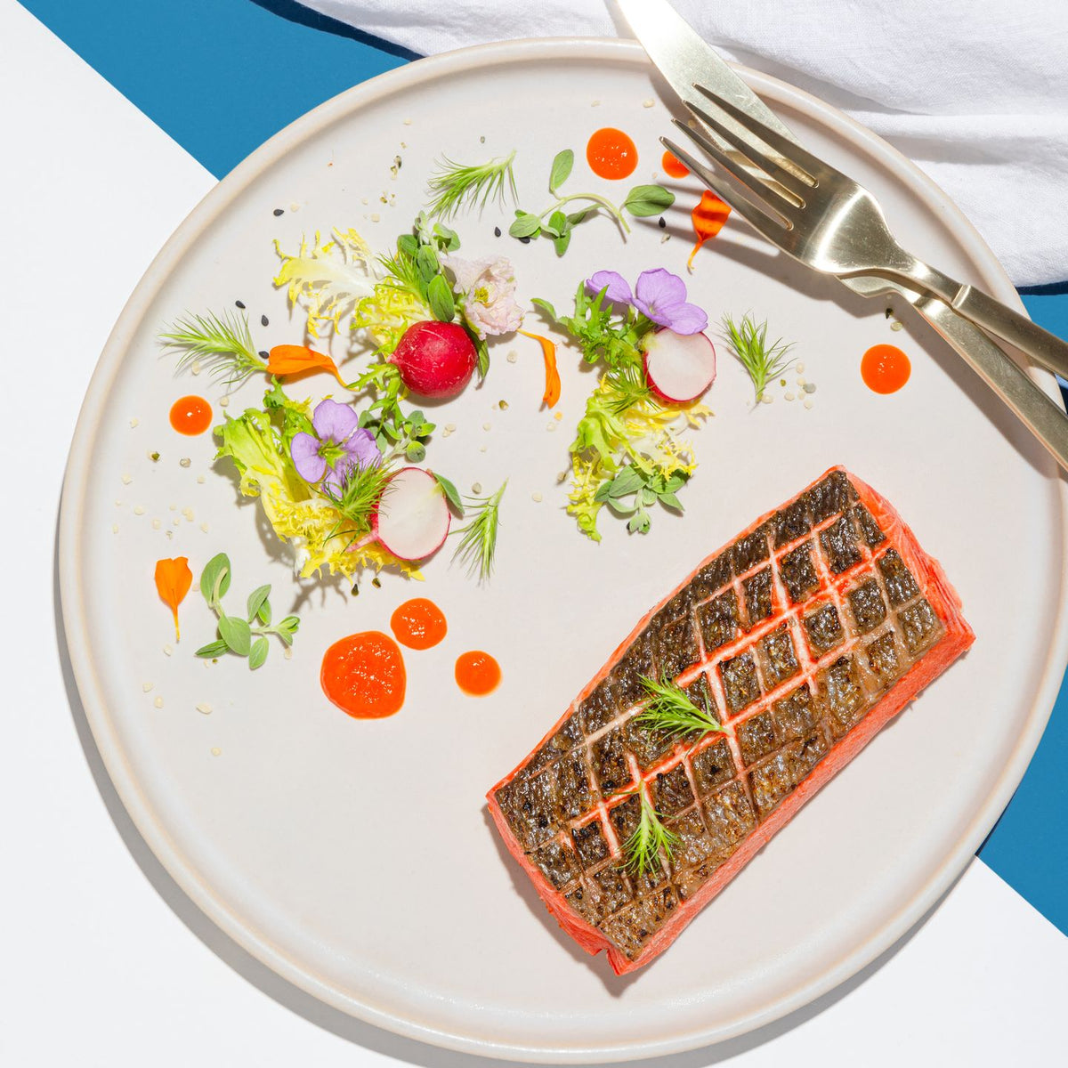 A seared salmon fillet with crosshatched crispy skin and a sprig of dill on a white plate, beside a small salad of radish, frisee and purple flowers, with gold cutlery and a linen napkin