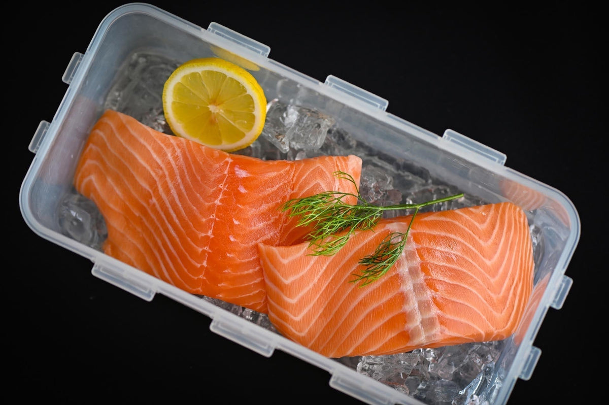 Top-down view of Alaskan salmon fillets showcasing bright orange flesh and white marbling on ice with lemon slice and dill