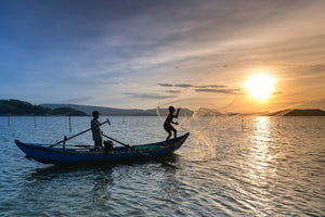 Two fishermen casting a circular net from a small boat at sunset on calm water