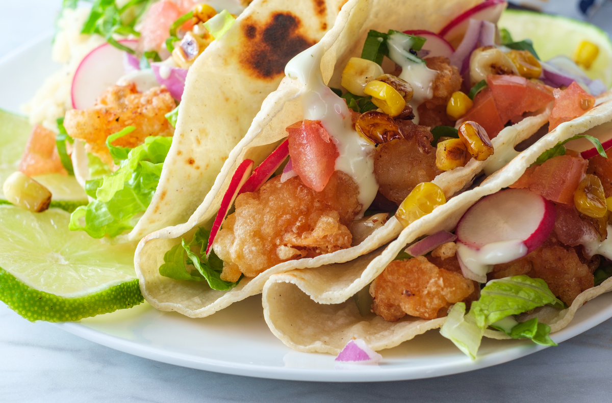 Close up of beer-battered salmon tacos with charred corn, radish slices, diced tomato, lettuce, crema, and lime wedges on soft tortillas