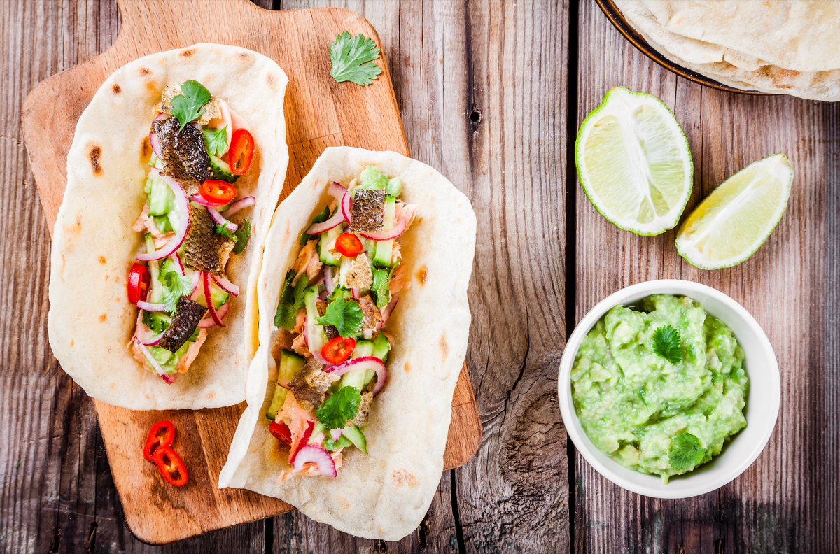 Two grilled salmon tacos on flatbreads with avocado guacamole and lime wedges on a wooden board.