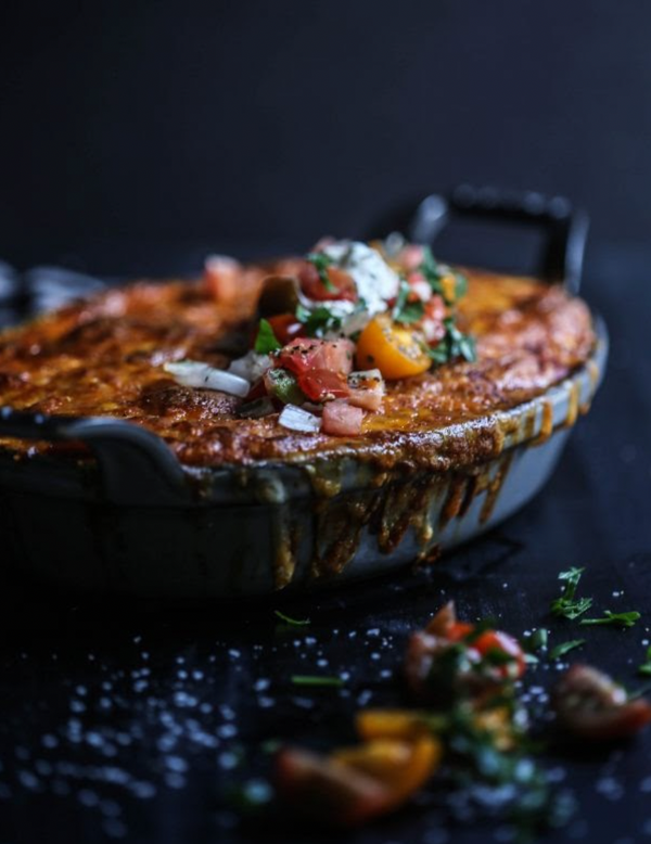 Skillet of cheesy baked enchiladas with browned, bubbling cheese and tomato-onion salsa garnish spilling down the side.