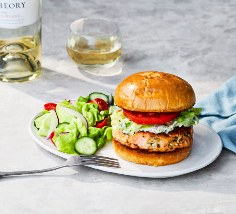 Salmon burger with creamy herb sauce and tomato on a brioche bun, with side salad and glass of white wine