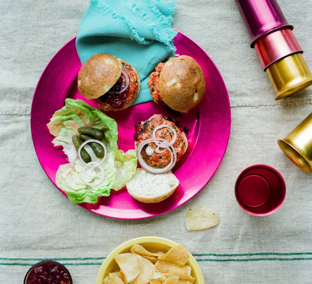 Two salmon burger sliders topped with rhubarb chutney and onion on a bright magenta plate, lettuce, cornichons and chips.