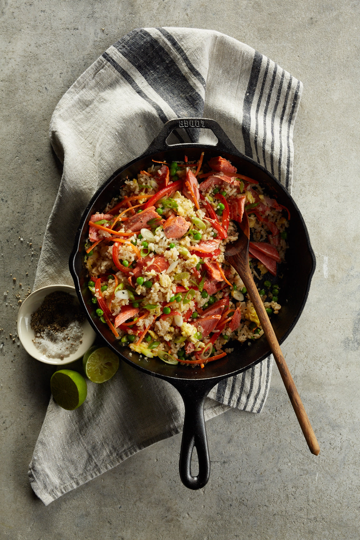 Cast-iron skillet of fried rice with flaked sockeye salmon, peas, red pepper and scallions; wooden spoon and lime halves