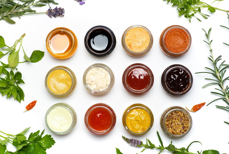 Twelve small glass bowls with different colored sauces arranged in rows, surrounded by fresh herbs and chili peppers on white background