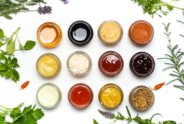 Twelve small glass bowls with different colored sauces arranged in rows, surrounded by fresh herbs and chili peppers on white background
