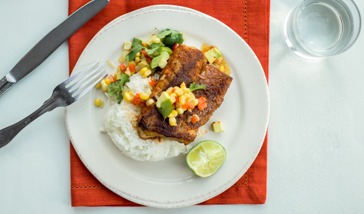 Blackened Wild Alaskan cod on rice with avocado corn salsa and lime on white plate over red napkin, silverware and water glass