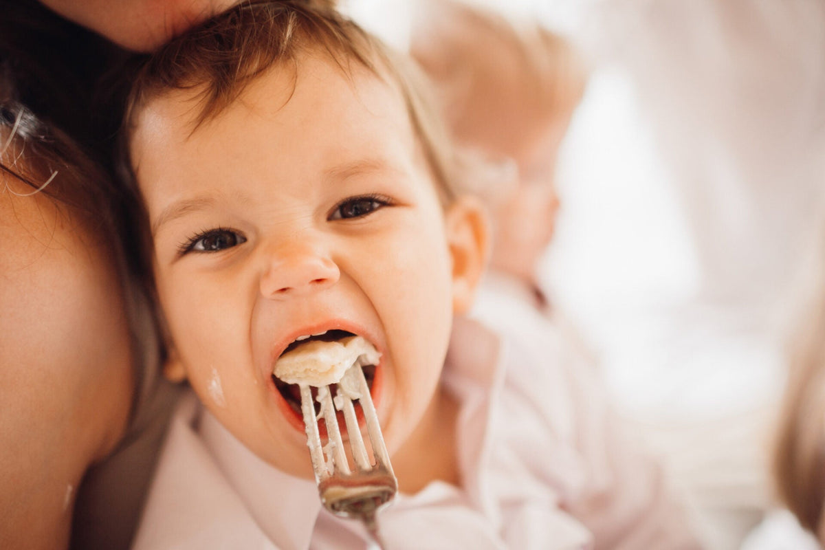 Close-up of a baby being fed a bite of mashed food from a fork while an adult holds them
