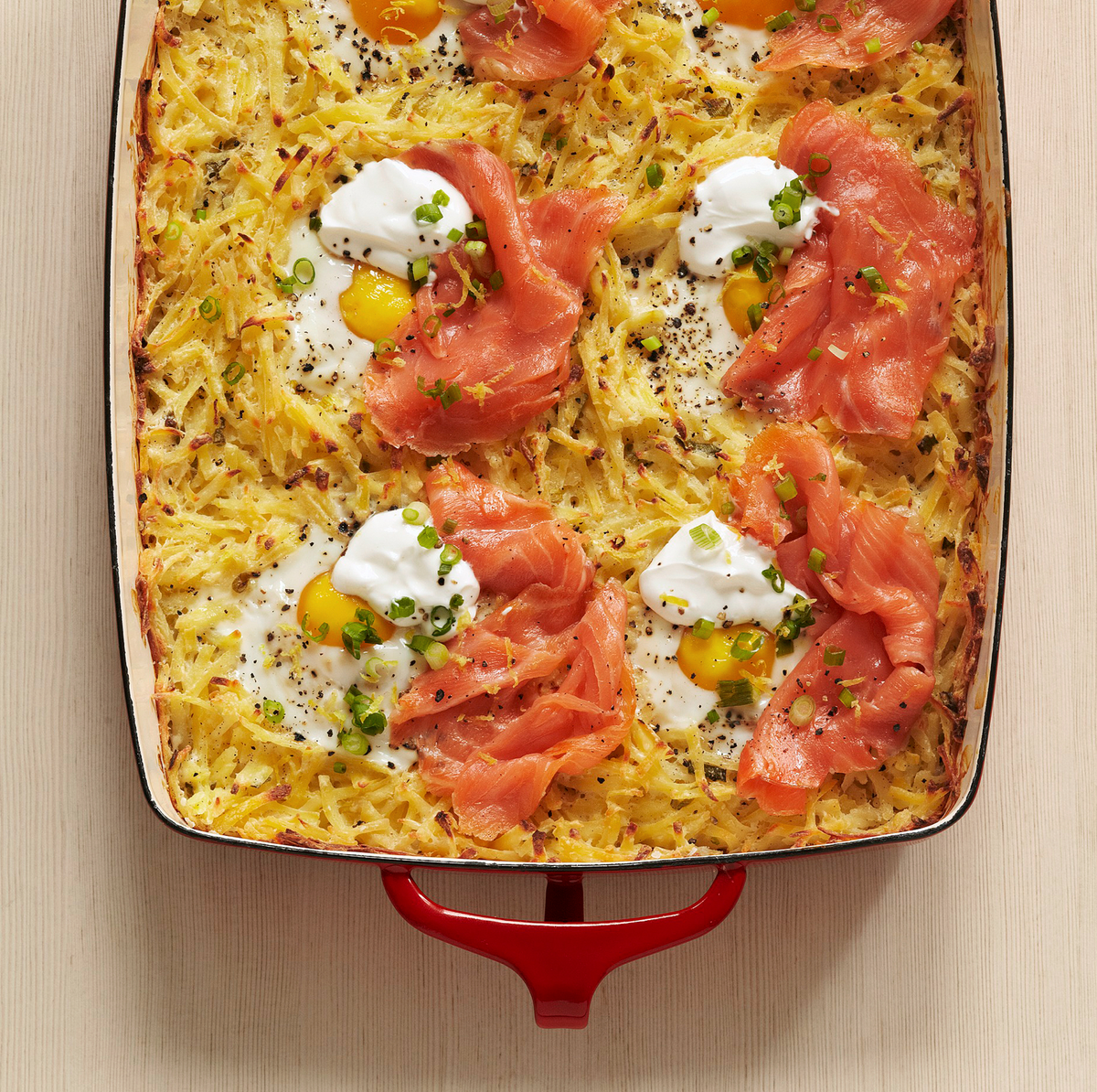 Baked hash brown casserole with eggs, smoked salmon, sour cream, and green onions in a red-handled pan