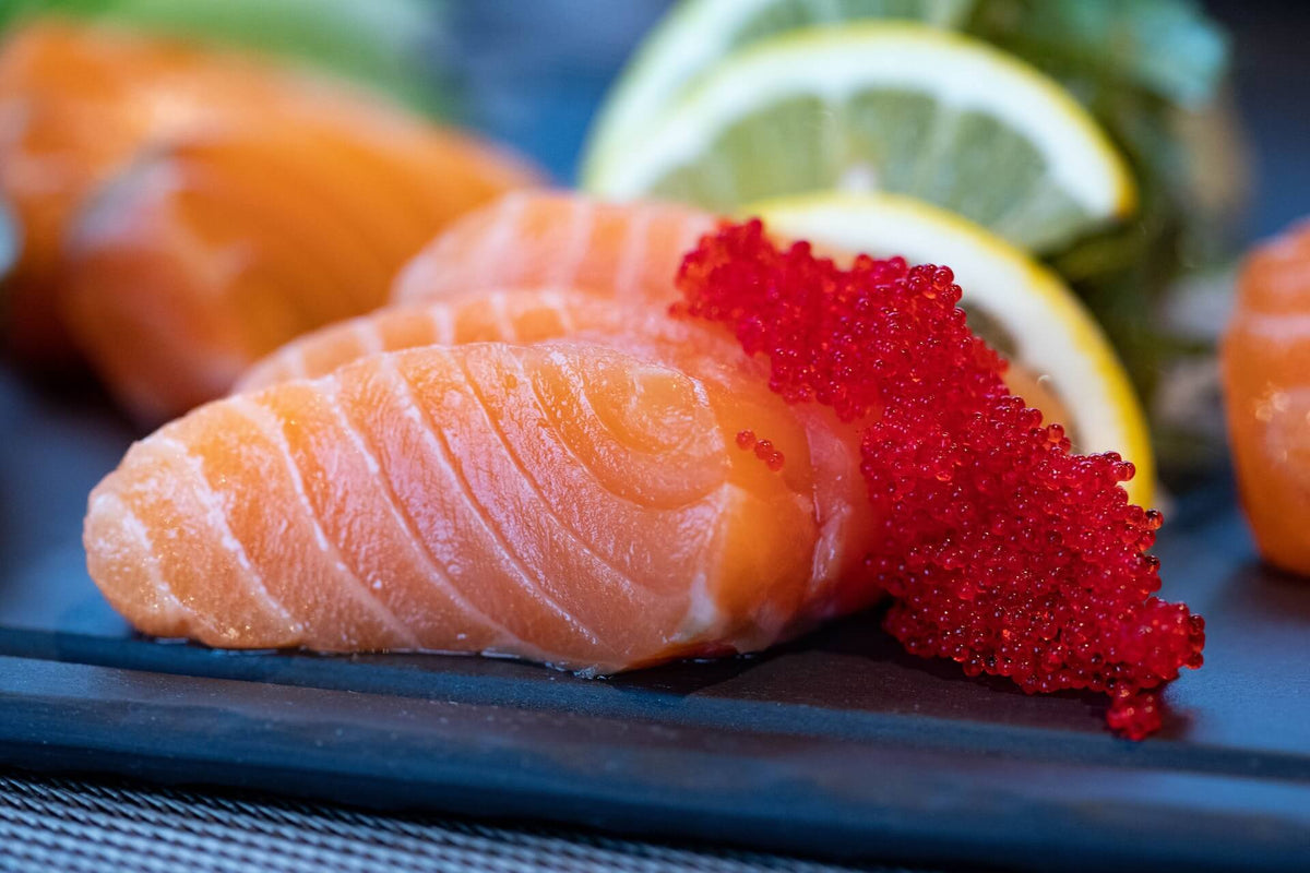 Close-up of raw salmon sashimi topped with red fish roe and lemon slices on a dark plate.