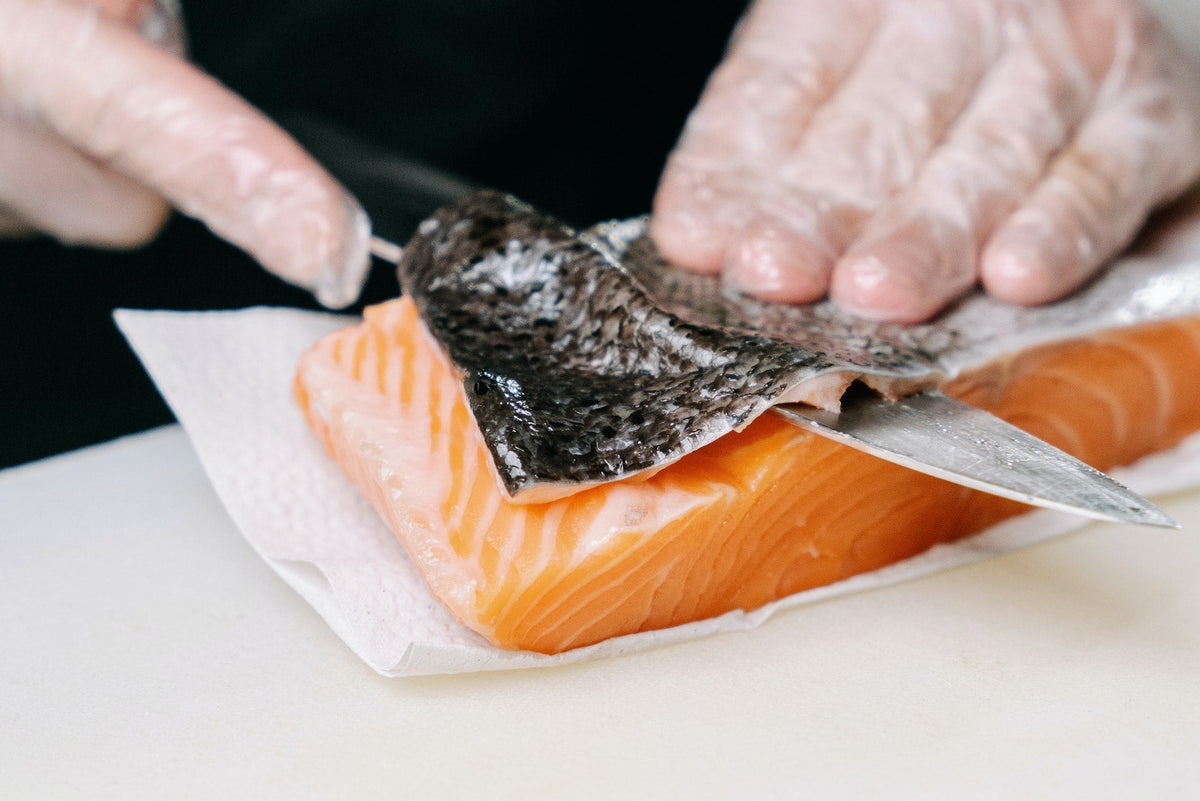 Hands using a fillet knife to remove skin from a raw Alaskan salmon fillet on a cutting board