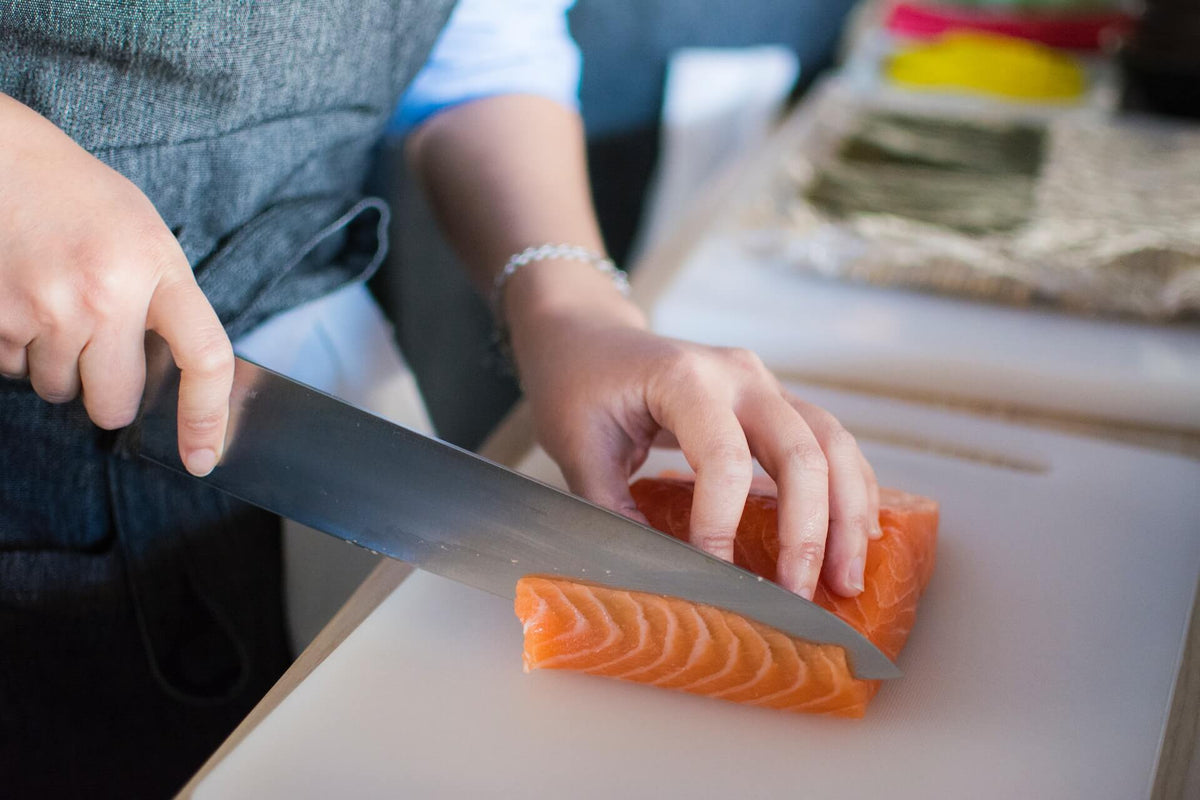 Hands slicing a raw salmon fillet with a chef's knife on a white cutting board in a kitchen