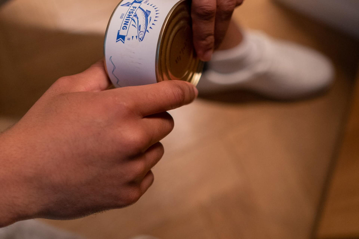 Close-up of a can of salmon showing blue 'FISHING Club' fish logo held in hands against a blurred hardwood floor