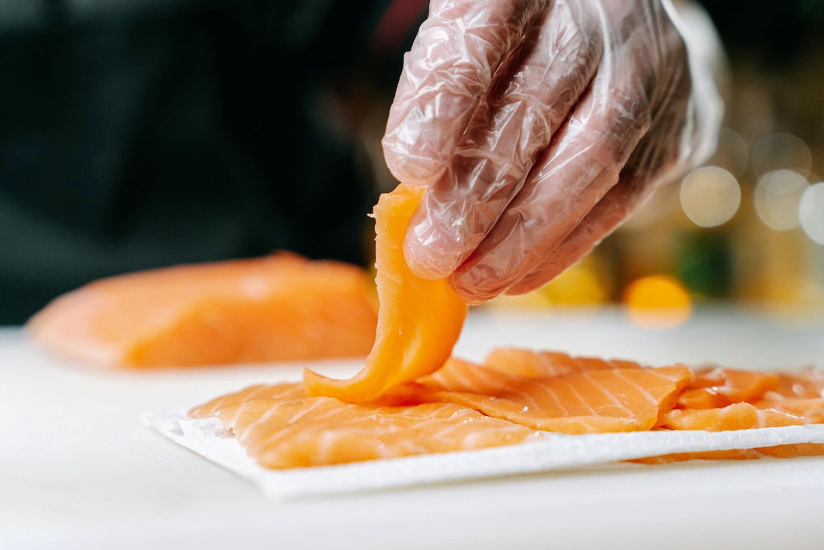 Gloved hand arranging thin slices of raw salmon on a paper towel