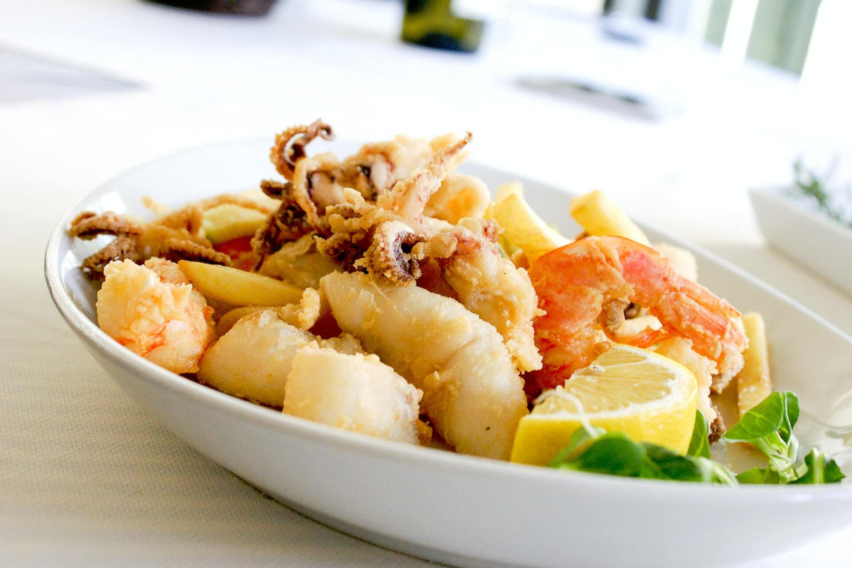 Close-up of fried mixed seafood (shrimp, calamari, battered fish) with lemon wedges and greens on a white ceramic plate