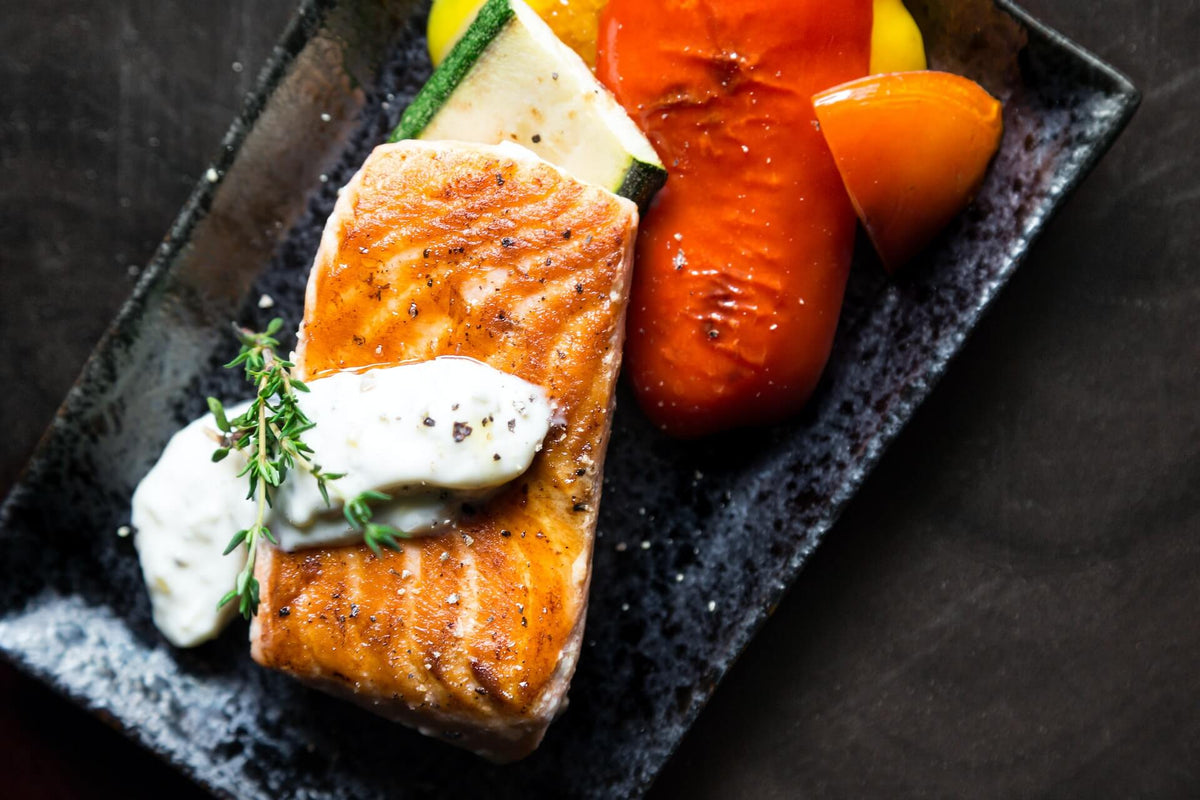 Pan-seared salmon fillet with herb cream sauce, grilled zucchini and tomato on a black rectangular plate.
