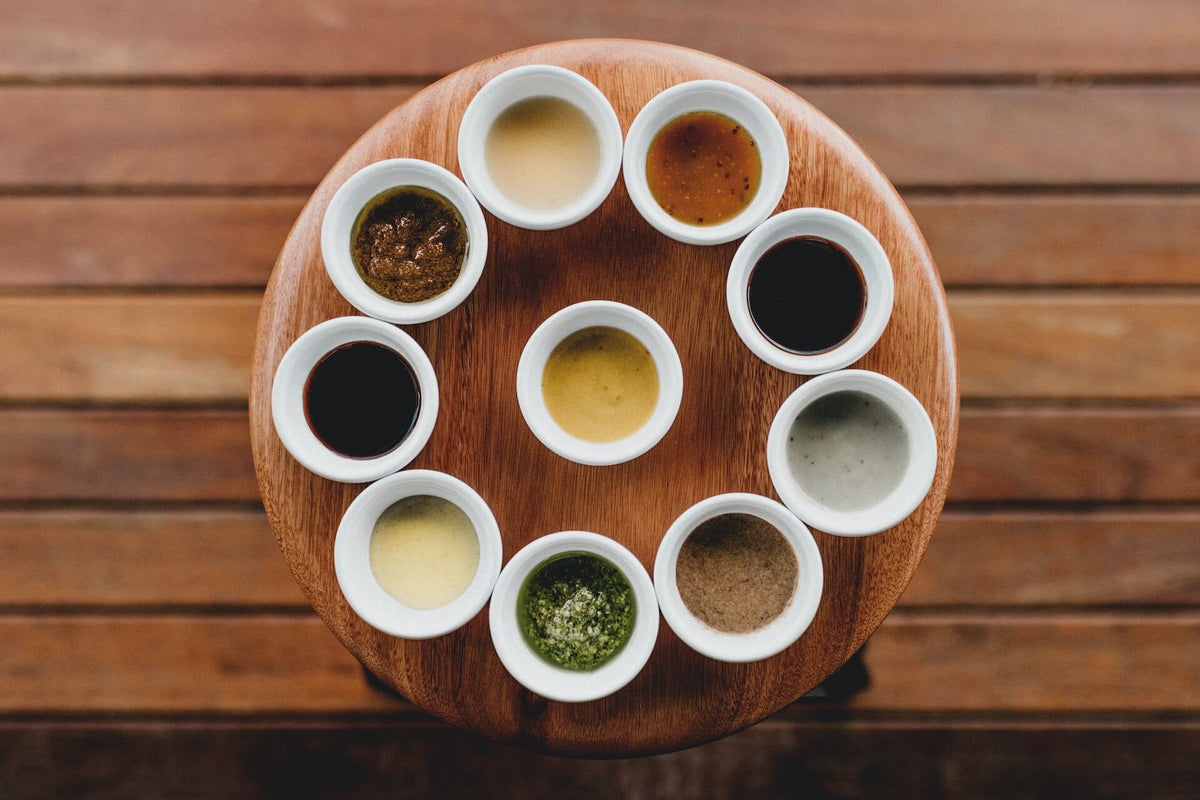 Ten small bowls of various sauces arranged in a circle on a round wooden table