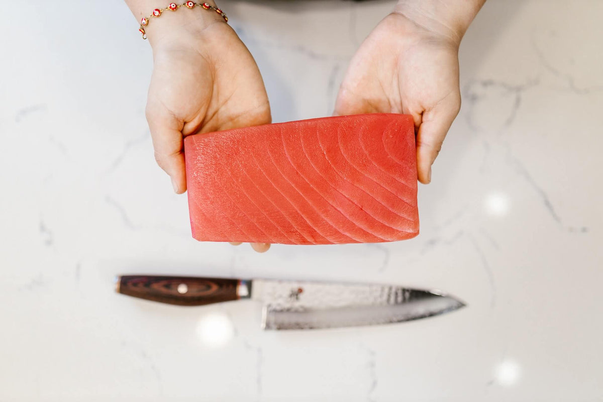 Raw tuna loin held in two hands above a chef's knife on a marble countertop