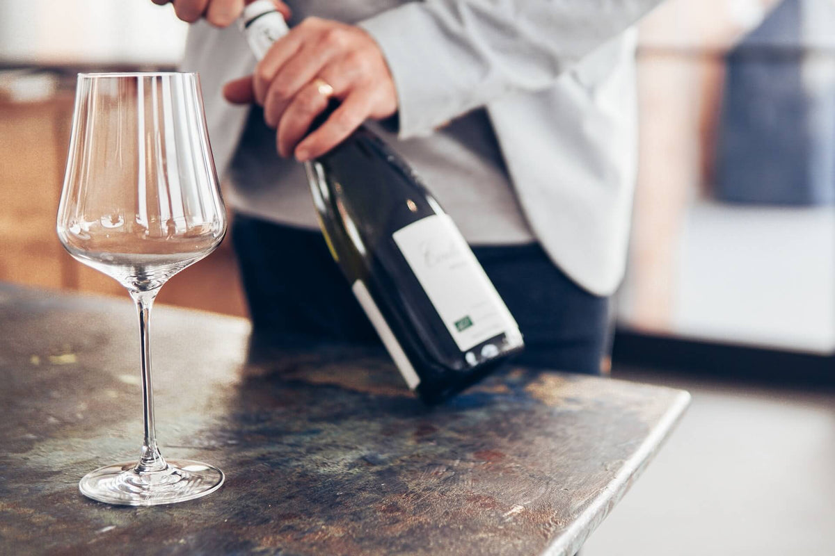 Person opening a wine bottle next to an empty wine glass on a table