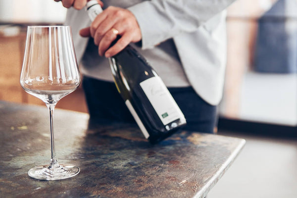 Person opening a wine bottle next to an empty wine glass on a table
