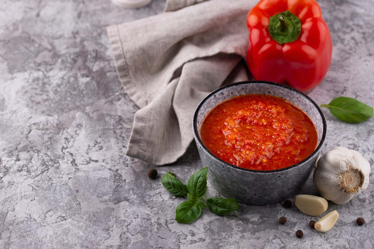 bowl of red marinade sauce with garlic cloves, basil leaves, and red bell pepper on gray surface