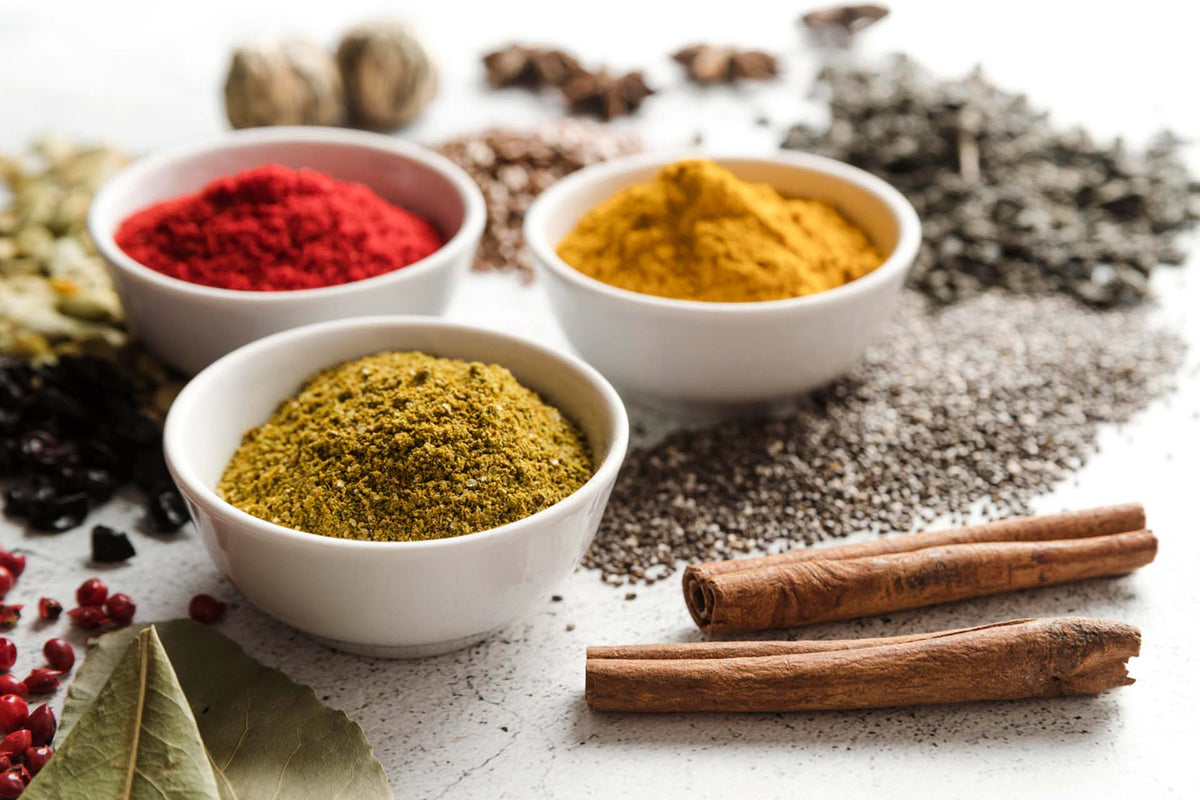 Three white bowls with red, yellow, and green spices, cinnamon sticks, bay leaves, and scattered seeds on a white surface