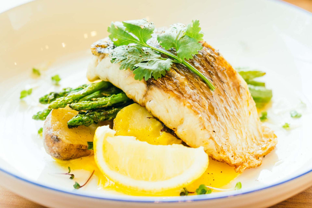 Cooked cod fillet with asparagus, potatoes, lemon wedge, and herbs on a white plate
