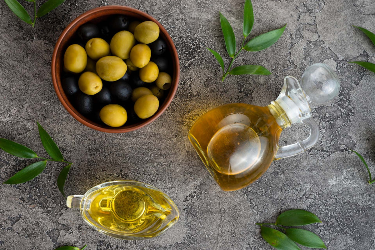 bowl of black and green olives with two glass containers of olive oil and green leaves on a gray textured surface