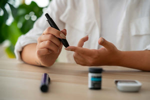 Close-up of hands using a lancing device to prick a fingertip with a glucose meter and test strip container on a table