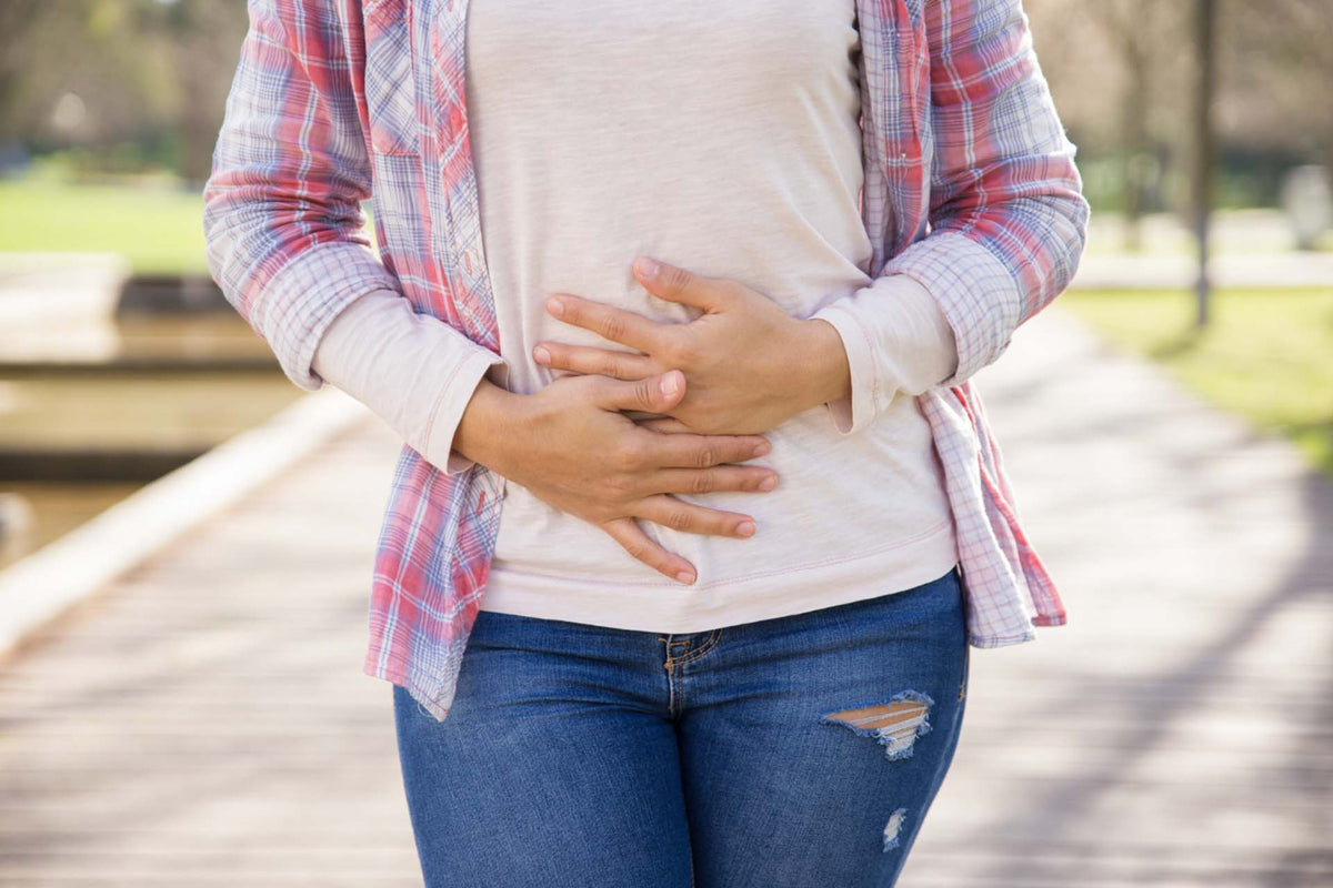 Woman clutching her lower abdomen on an outdoor boardwalk, midsection view suggesting abdominal discomfort