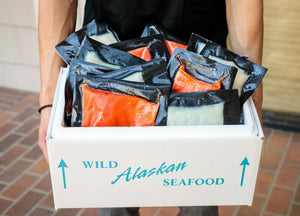 Box labeled 'Wild Alaskan Seafood' filled with vacuum-sealed packages of red and white fish held by a person.