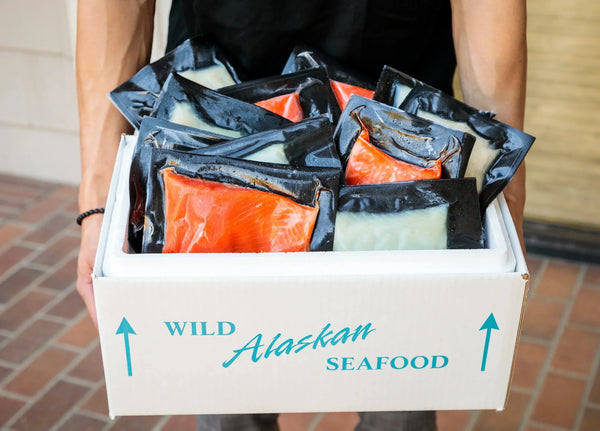 Box labeled 'Wild Alaskan Seafood' filled with vacuum-sealed packages of red and white fish held by a person.