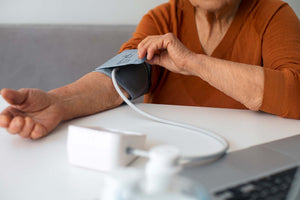 Older woman measuring her blood pressure with a home arm cuff while seated at a table.