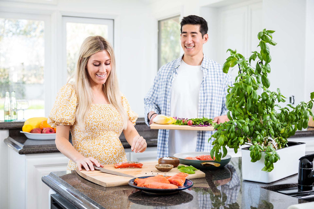 Pregnant woman seasoning wild sockeye salmon fillets on a kitchen island as her partner smiles and holds a tray of salad greens