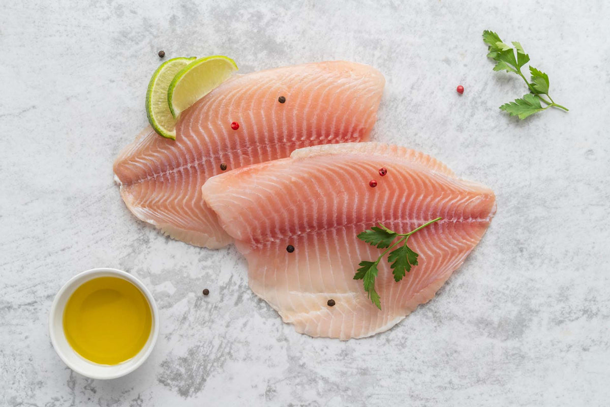 Top-down view of two raw tilapia fillets with pale pink flesh, lime wedges, parsley, and olive oil on a light stone surface