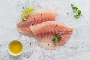 Top-down view of two raw tilapia fillets with pale pink flesh, lime wedges, parsley, and olive oil on a light stone surface