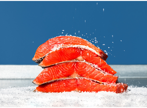 Three stacked raw sockeye salmon fillets sprinkled with coarse salt on a white surface and blue background.