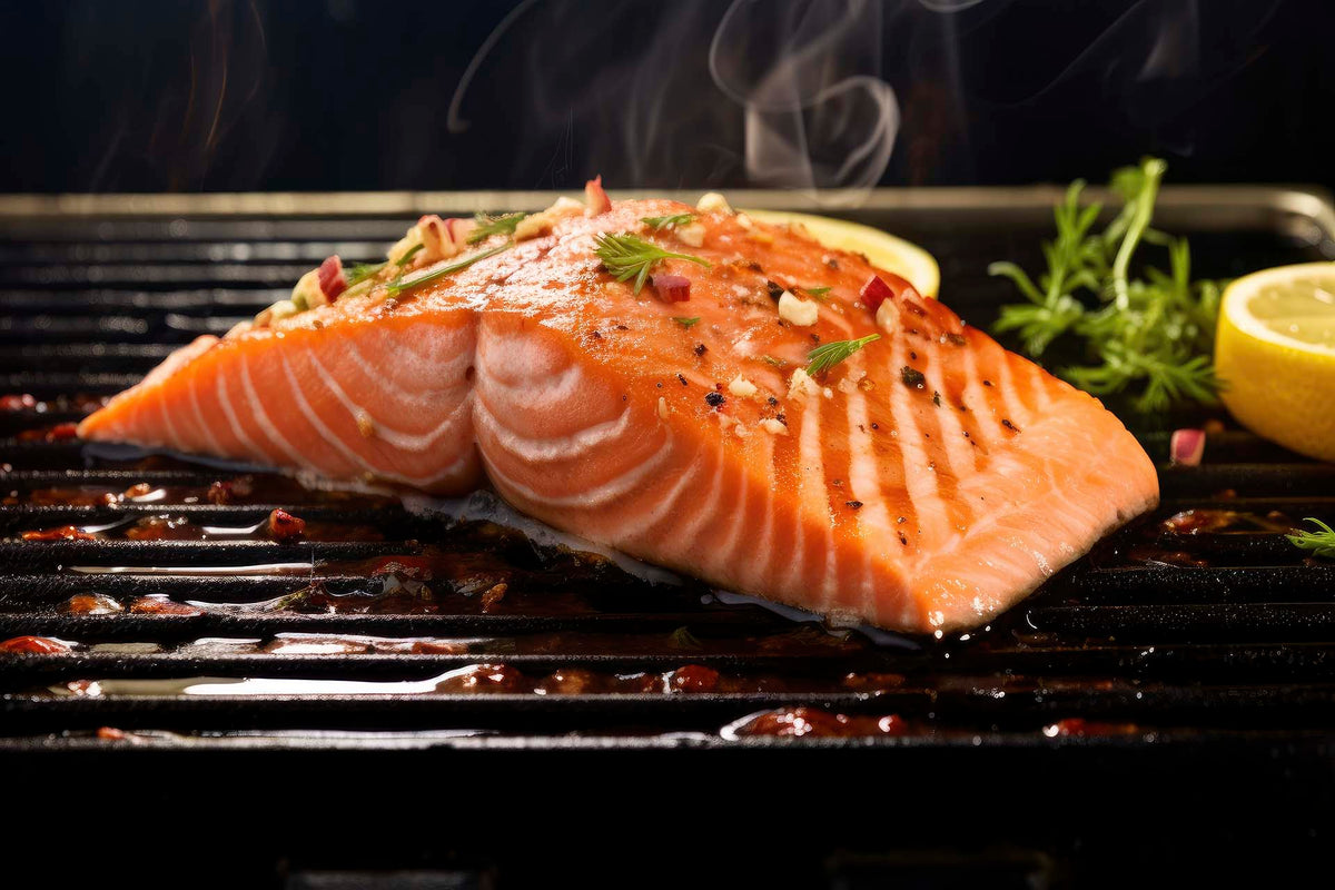 Close-up of Alaskan salmon fillet on a hot grill showcasing charred grill marks, chopped herbs and garlic, and lemon halves with steam rising