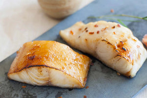 Close up of two seared Alaskan black cod fillets showcasing golden glazed tops and flaky white flesh on a dark slate plate