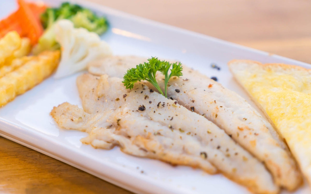 Close-up of cooked white fish fillets showing flaky texture and cracked black pepper on a white plate with vegetables and buttered toast