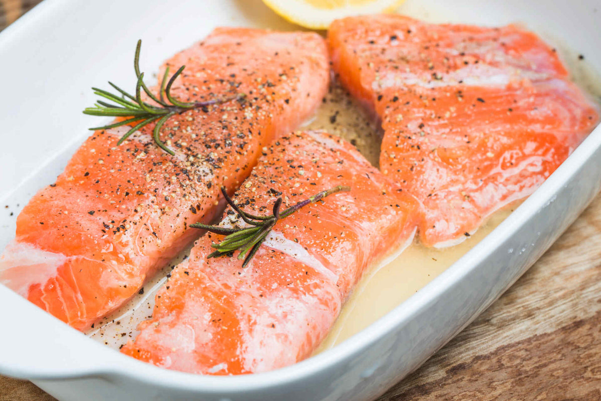 Close-up of salmon fillets marinating in a white dish, seasoned with cracked pepper and rosemary, lemon wedge visible