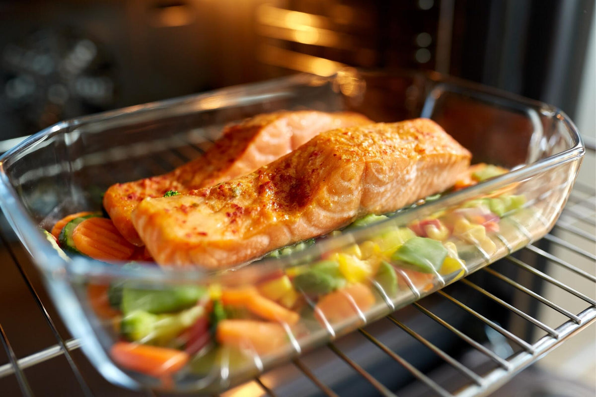 Two seasoned salmon fillets on mixed vegetables in a glass baking dish inside an oven