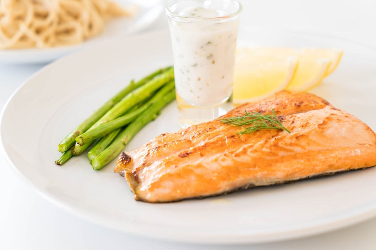 Grilled salmon fillet with asparagus, lemon wedges and a small glass of creamy dill sauce on a white plate.