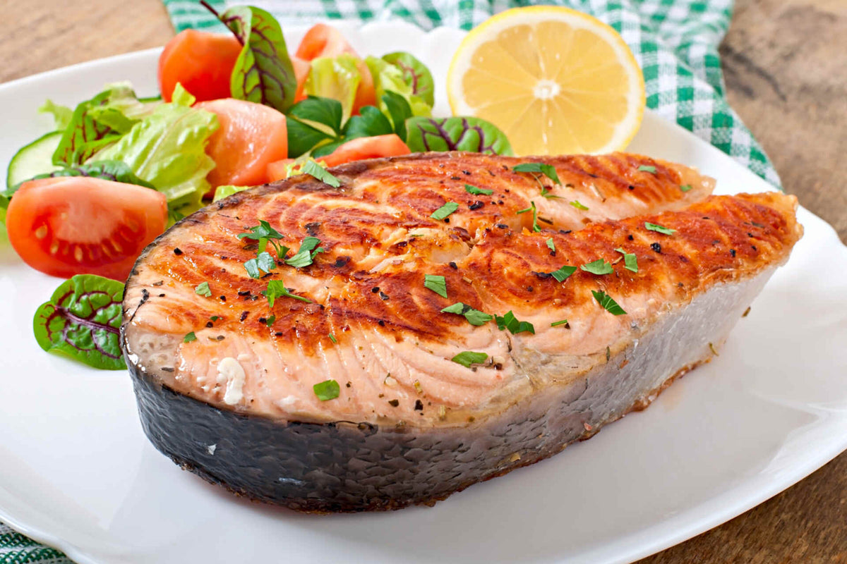 Close-up of grilled Alaskan sockeye salmon steak showing charred grill marks and parsley garnish on a white plate with salad and a lemon half