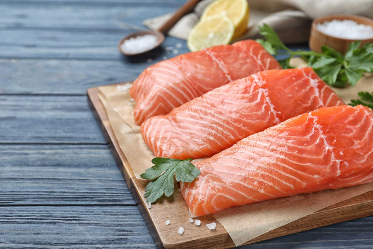Close-up of three raw Alaskan salmon fillets showing bright orange flesh and white marbling on a wooden board with parsley, lemon, and coarse salt