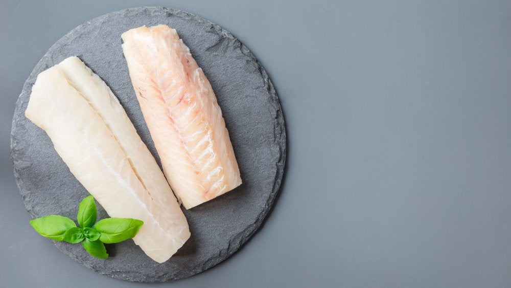 Two raw white fish fillets on a round slate board with a small basil sprig