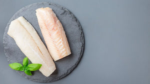 Two raw white fish fillets on a round slate board with a small basil sprig