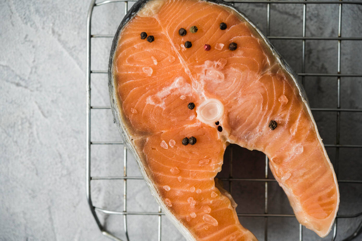 Overhead view of raw salmon steak seasoned with coarse salt and whole peppercorns on a metal cooling rack